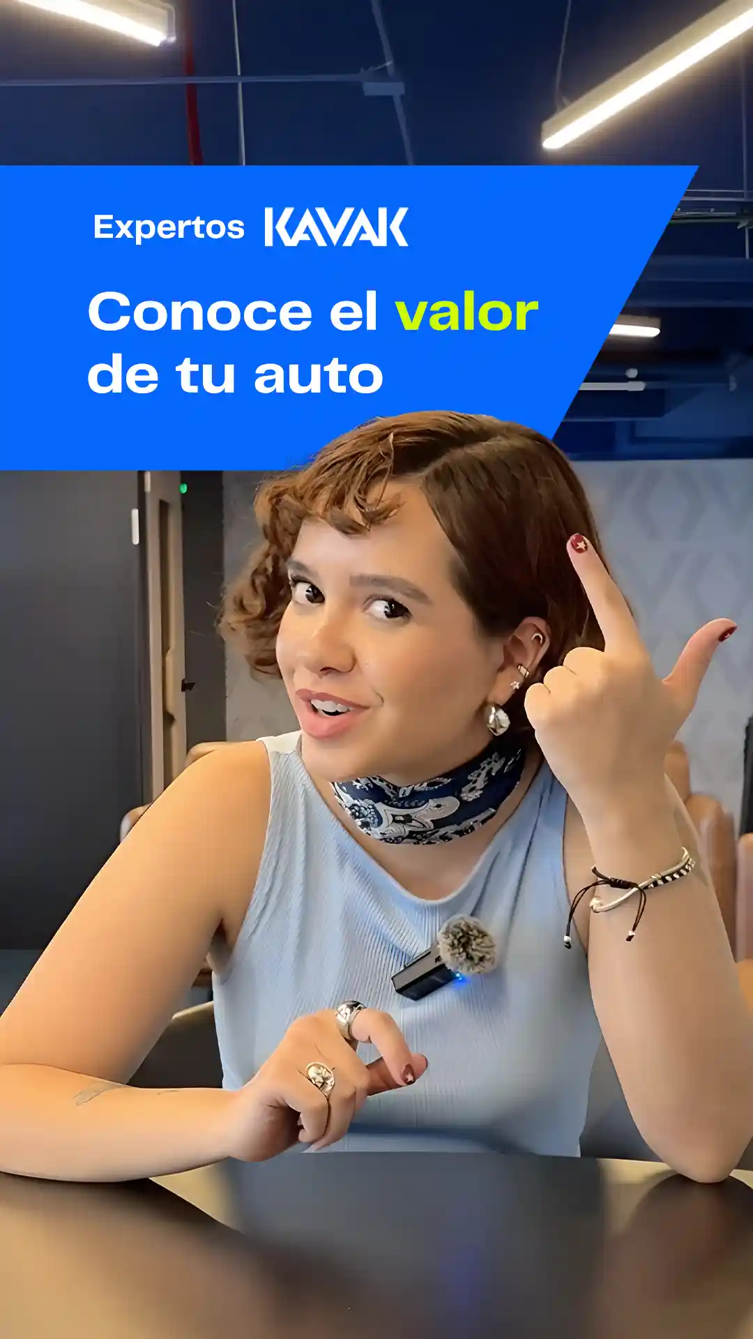 Mujer joven con blusa azul claro y mascada azul, sonriendo mientras señala su cabeza con una mano y sostiene un micrófono con la otra.