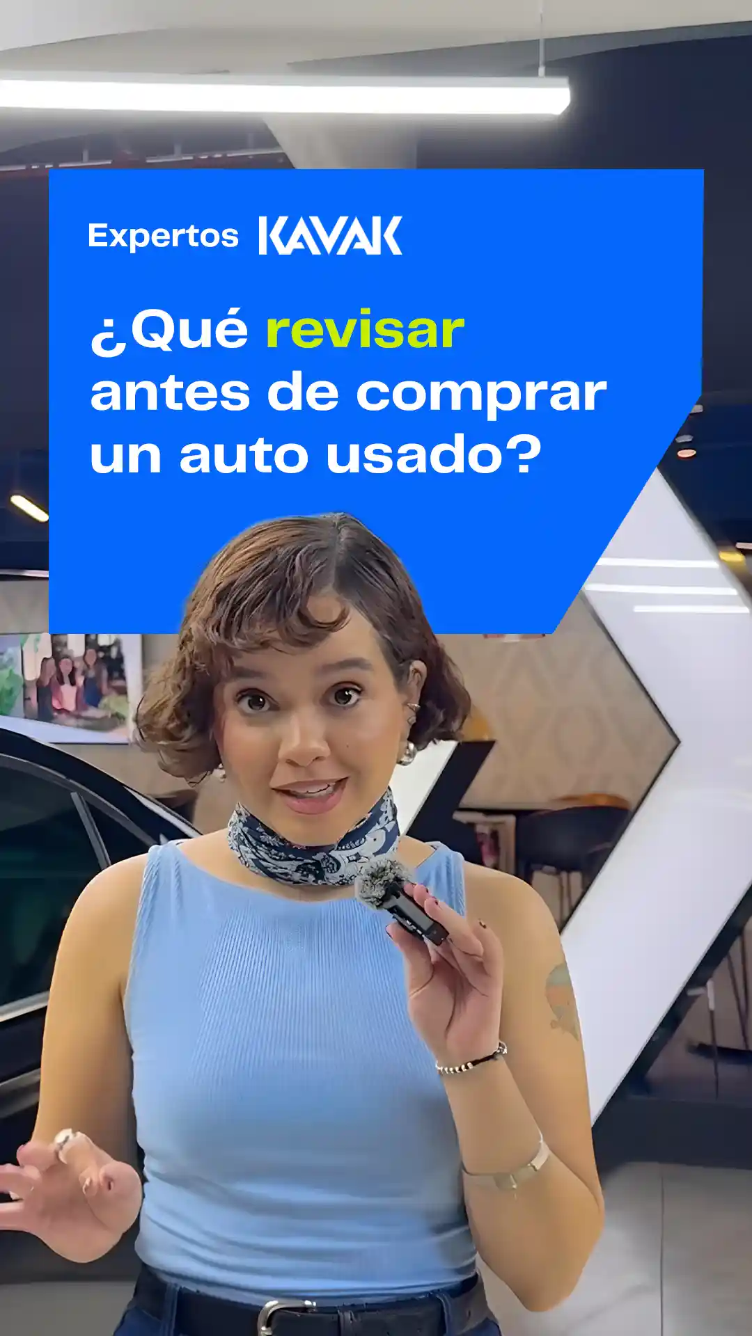 Mujer joven con blusa azul claro y mascada, hablando a cámara con micrófono en mano, frente a un auto.